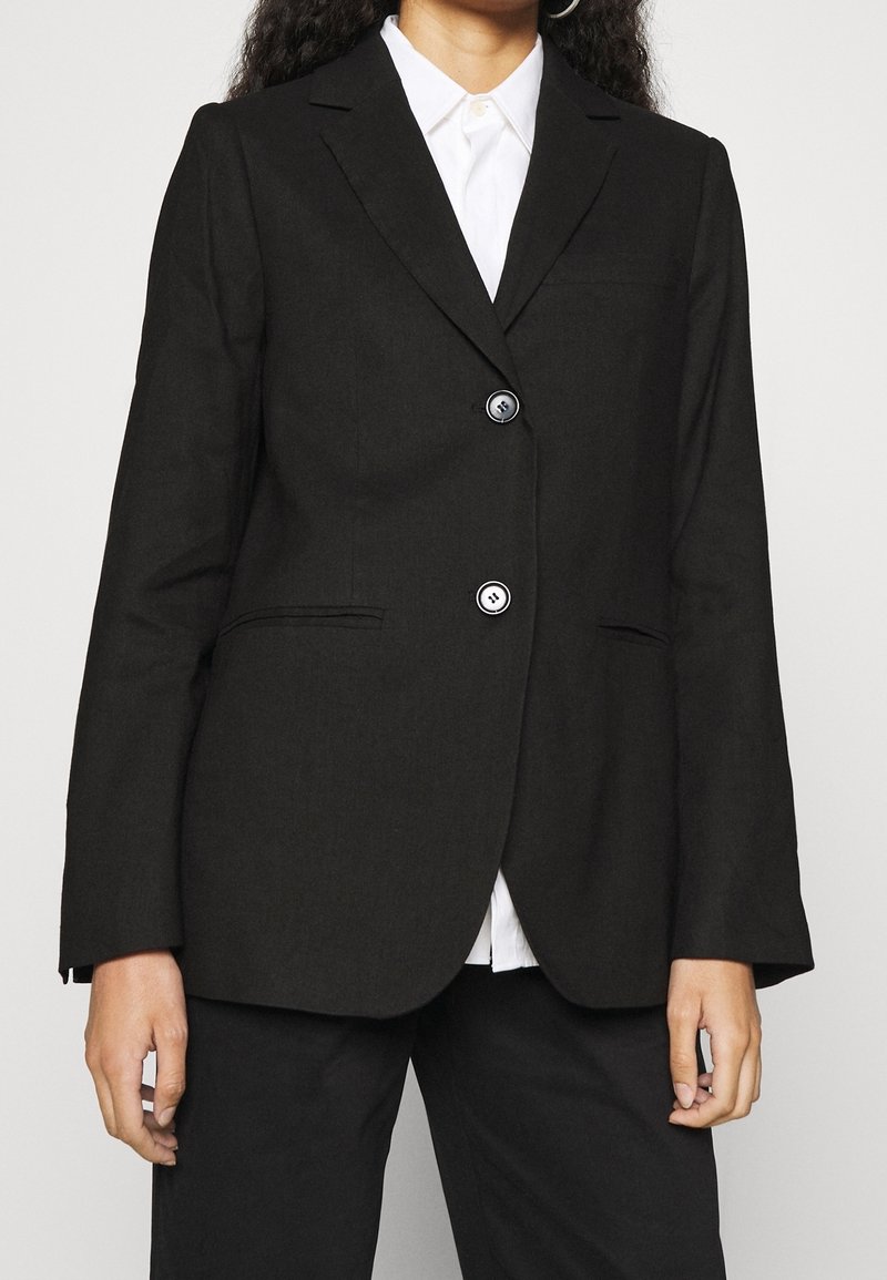 Blazer noir à coupe ajustée, avec deux boutons, col à revers crantés et poches latérales. Le tissu présente une texture lisse et structurée.