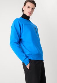 Felpa blu realizzata in tessuto morbido, con tasca con zip sul petto e polsini a costine, abbinata a pantaloni neri.