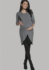 Graues gestricktes Schwangerschafts Kleid mit Dreiviertelärmeln, asymmetrischem Saum, geschichteter Optik, kombiniert mit schwarzen Strumpfhosen und knöchelhohen Stiefeln mit Absatz.