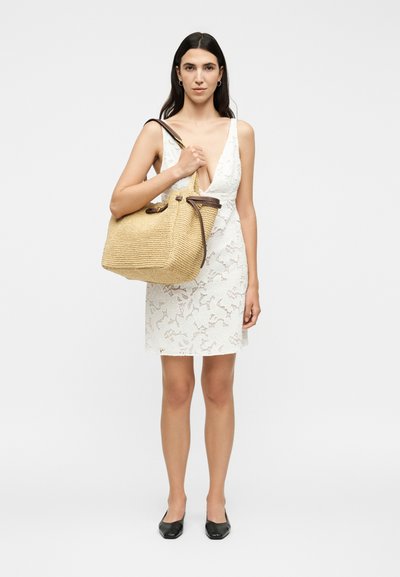 Γυναίκα με λευκό δαντελένιο φόρεμα χωρίς μανίκια, κρατώντας μεγάλη μπεζ υφαντή τσάντα με καφέ δερμάτινες λαβές, φορώντας μαύρες μπαλαρίνες σε απλό φόντο.