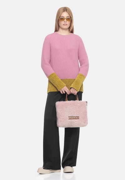 Maglione rosa a coste con dettagli in oro, abbinato a pantaloni neri larghi e una borsa rosa pelosa. La modella indossa occhiali da sole gialli.