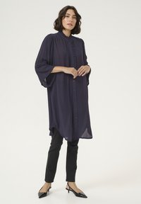 Chemise boutonnée longue de couleur violet foncé avec un devant plissé et des manches larges, assortie à un pantalon noir ajusté et des escarpins noirs à bout pointu.