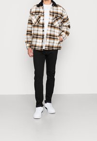 Person i vit t-shirt, brun och svart rutiga jacka, svarta byxor och vita sneakers som står framför en jämn vit bakgrund.