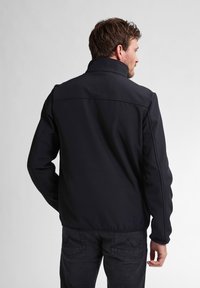 Giacca softshell nera con colletto alto, caratterizzata da una texture liscia e un design elegante. Indossata con jeans scuri, mostra una silhouette moderna.