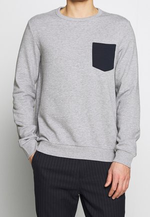 Mand iført lysegrå langærmet sweatshirt med mørk brystlomme og sorte bukser med nålestriber, hænder i lommerne, mod ensfarvet baggrund.