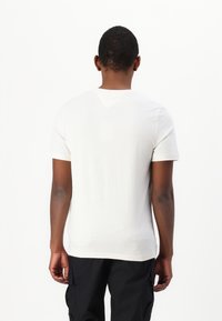 Camiseta de algodón blanca de manga corta, con cuello redondo y un diseño liso, que se lleva con pantalones cargo negros, vista desde la parte de atrás.