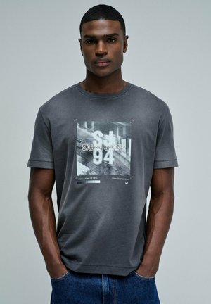 Hombre negro con cabello corto que lleva una camiseta gris con el gráfico "Salsa Jeans 94" y vaqueros azules, de pie con las manos en los bolsillos sobre un fondo liso.