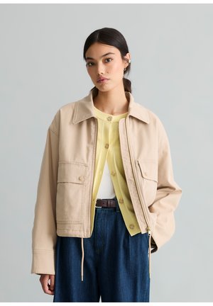 Junge Frau mit dunklem Haar trägt eine beige Jacke über einem gelben Cardigan, in hoch taillierte dunkelblaue Jeans gesteckt, stehend vor einem grauen Hintergrund.