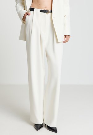 Personne portant un pantalon blanc taille haute à jambes larges avec ceinture noire, un blazer blanc, les mains dans les poches, et des talons noirs à bouts pointus.