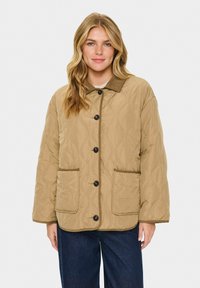 Quiltad beige jacka med krage, knappstängning och två framfickor. Har en strukturerad yta och kontrasterande trim.