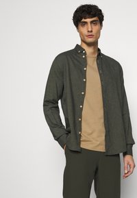 Olivgrünes Hemd mit Knöpfen in hellbraun, getragen über einem beigen T-Shirt, kombiniert mit olivgrünen Hosen; glatte Textur und lässiges Design.