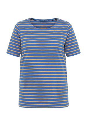 Femme aux longs cheveux ondulés portant un t-shirt à manches courtes rayé bleu et orange ainsi qu’un pantalon bleu clair, debout contre un mur blanc.