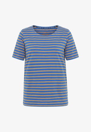 Frau mit langen, welligen Haaren, die ein blau-orange gestreiftes Kurzarmshirt und hellblaue Hose trägt, steht an einer weißen Wand.