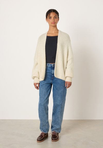 Cardigan en grosse maille crème surdimensionné avec poignets côtelés, associé à un jean ample bleu clair et des chaussures marron à lacets. Style simple, sans motifs.