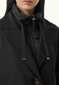 Cappotto nero a strati con giacca interna imbottita, colletto alto, dettagli con cerniera e cordini regolabili. Accento di bottone notevole.