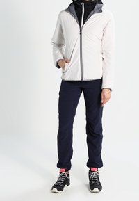 Veste zippée gris clair avec capuche, associée à un pantalon bleu marine et des baskets noires. La veste présente une texture lisse et des accents contrastés.
