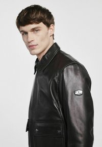 Czarna skórzana kurtka z wywiniętym kołnierzem, z przodu zapinana na zamek, z bocznymi kieszeniami i charakterystycznym srebrnym logo na lewym rękawie. Gładka faktura.