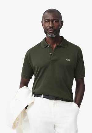 Polo shirt verde scuro con colletto classico, tre bottoni e logo Lacoste. Il modello tiene in mano una giacca bianca e indossa pantaloni chiari.