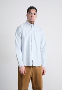 Light blue button-up shirt with a collar, featuring a pocket. Worn with brown corduroy pants, highlighting a relaxed fit and texture.
