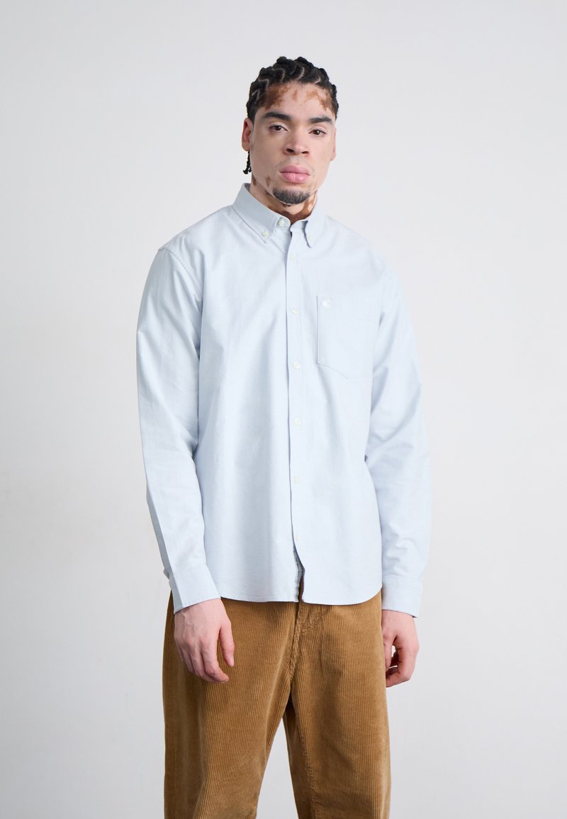 Light blue button-up shirt with a collar, featuring a pocket. Worn with brown corduroy pants, highlighting a relaxed fit and texture.