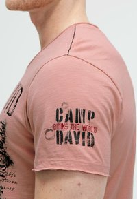 Light pink short-sleeve T-shirt with a crew neck, featuring distressed black and red text graphics, and raw edges on the sleeves and hem.