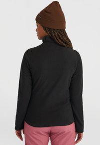 Zwarte langslavige shirt met hoge kraag, zachte textuur. Gemaakt met een bruine beanie en roze broek, met een aansluitend ontwerp dat van achteren zichtbaar is.