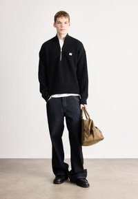 Čierny pletený pulóver s polovičným zipsom, v kombinácii s tmavými džínami. V jednej ruke je držaná hnedá kožená taška. Hladká textúra a voľný strih.
