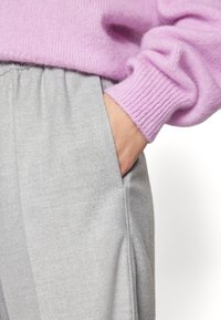 Personne portant un pantalon gris avec la main dans la poche et un pull rose avec un poignet côtelé visible au poignet.