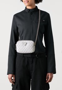 Crossbody torba svijetlo sive boje s geometrijskim uzorkom, logom i metalnom lancem; nosi se preko crne jakne i hlača.