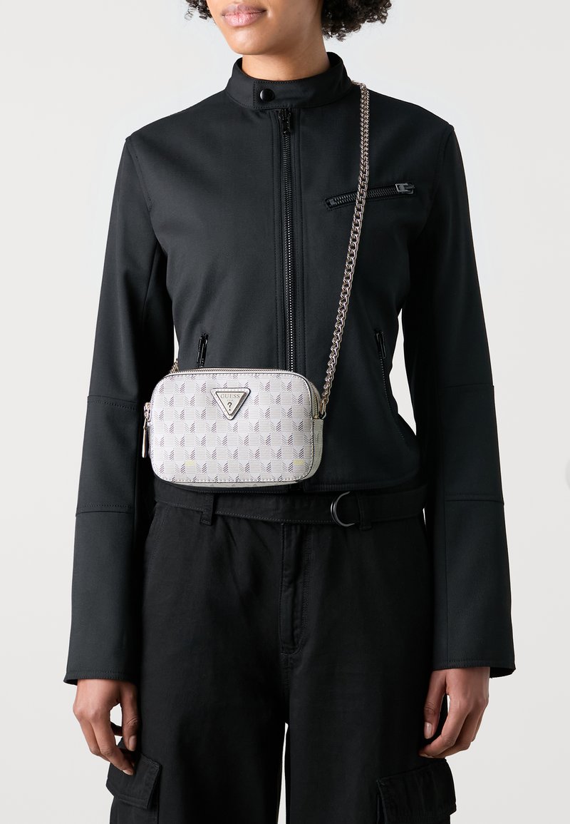 Crossbody torba svijetlo sive boje s geometrijskim uzorkom, logom i metalnom lancem; nosi se preko crne jakne i hlača.