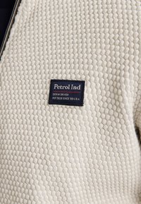 Texturovaná krémová bunda na zip s včelí pláství, se modrou obdélníkovou nálepkou s nápisem "Petrol Ind" a dalšími detaily níže.