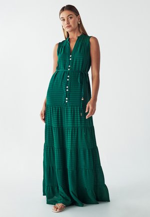 Green, sleeveless maxi dress made of textured fabric with tiered layers, button front, and a tied waist. Flat sandals visible at the bottom.