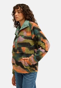 Femme aux cheveux bouclés porte une veste polaire colorée à motifs et un jean bleu, debout avec une main dans la poche de la veste, regardant sur le côté.