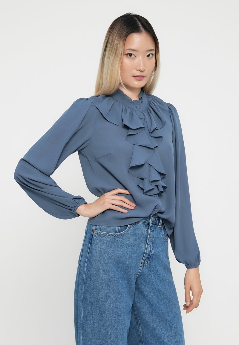 Blaue Rüschbluse mit hohem Kragen und Knopfverschluss, lange Ärmel und elastische Bündchen, kombiniert mit weit geschnittenen Jeans.