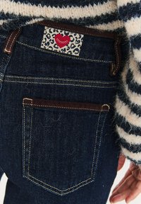 Tmavě modré džínové kalhoty s hnědou koženou nášivkou na pasu ozdobenou leopardím vzorem a červeným srdcem. Akcentované zlatým prošitím a zadními kapsami.