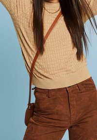 Žena v béžovém texturovaném svetru, hnědých semišových kalhotách, zlatém řetízku a s hnědou crossbody kabelkou na modrém pozadí.