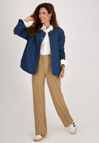 Gequiltede marineblauwe jas met gouden drukknoopjes, witte blouse, en beige wijde broek; gecombineerd met witte sneakers met zwarte strepen.