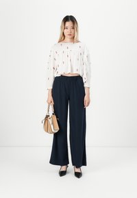 Woman with long hair wearing a white floral blouse, navy wide-leg pants, black heels, and holding a tan handbag with chain handles.
