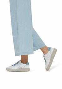 Weiße Sneakers mit hellblauen Akzenten, strukturierten Gummisohlen und flachen Schnürsenkeln. Kombiniert mit weiten, hellblauen Jeans.