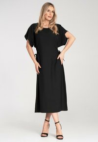 Schwarzes Midi-Kleid mit weiten Ärmeln, seitlichen Bindebändern und lockerem Schnitt. Glattes Material mit minimaler Textur. Getragen mit schwarzen Riemchen-Heels.