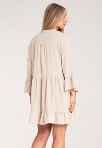 Beżowa sukienka z warstwowym wzorem, posiadająca rozszerzane rękawy, teksturowany materiał i falbaniasty dół. Tylny widok ukazuje plisowane detale na ramionach.