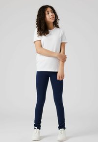 T-shirt en coton blanc associé à des leggings navy, présentant un design ajusté. La personne porte des baskets blanches et a les cheveux bouclés.