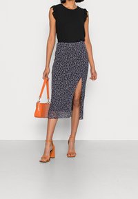 Donna che indossa un top senza maniche nero, una gonna midi fiorata blu marina con spacco frontale, mentre tiene una borsa arancione e indossa tacchi arancioni coordinati.