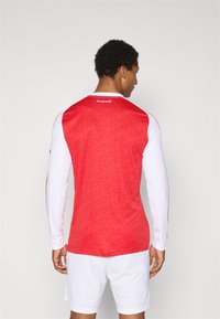 Rød og hvid langærmet sports trøje med et tekstureret rødt mønster, hvide ærmer og "Arsenal" logo på den øverste ryg.