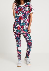 Kvinna som bär en färgglad grafisk t-shirt och matchande leggings med tecknade ögon, blommor och abstrakta former, tillsammans med vita sneakers.