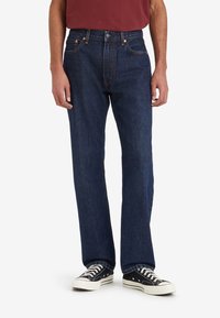 Mörkblå denimjeans med rak ben-design, med orange sömmar, knappfäste och femficksstil, kombinerade med svarta sneakers.