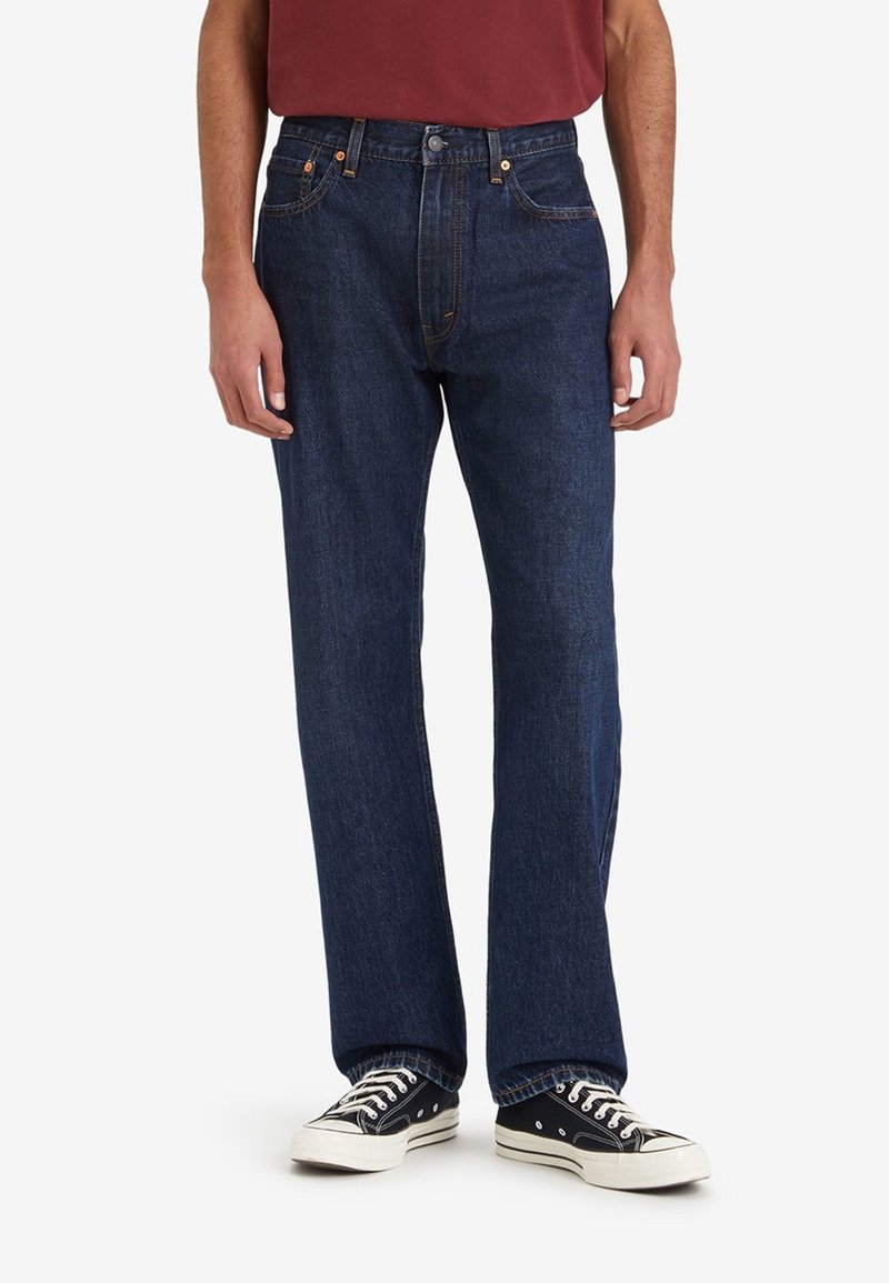 Mörkblå denimjeans med rak ben-design, med orange sömmar, knappfäste och femficksstil, kombinerade med svarta sneakers.