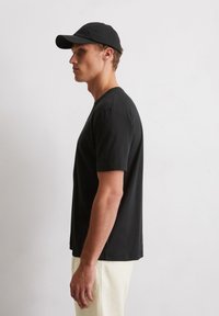 Zwarte katoenen t-shirt met korte mouwen en een ronde hals, gecombineerd met een zwarte pet, gepresenteerd tegen een neutrale achtergrond.