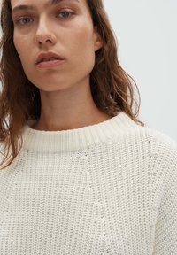 Pull en maille crème avec un col rond côtelé et une couture texturée distincte. Le tissu semble doux et léger avec une coupe ample.