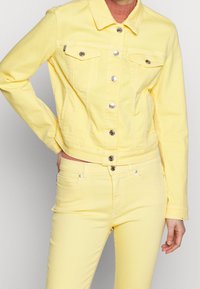 Giacca di denim gialla con bottoni argentati, due tasche sul petto e design corto. Abbinata a jeans in denim giallo coordinati.